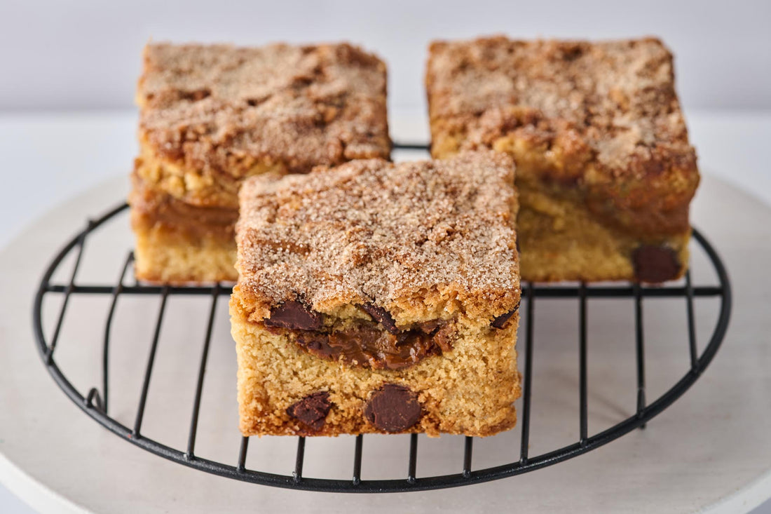 Churro Dulce de Leche Blondie with buttery cinnamon blondie base, swirled with dulce de leche, Ghirardelli dark chocolate chips, and topped with cinnamon sugar for a rich, caramelized treat