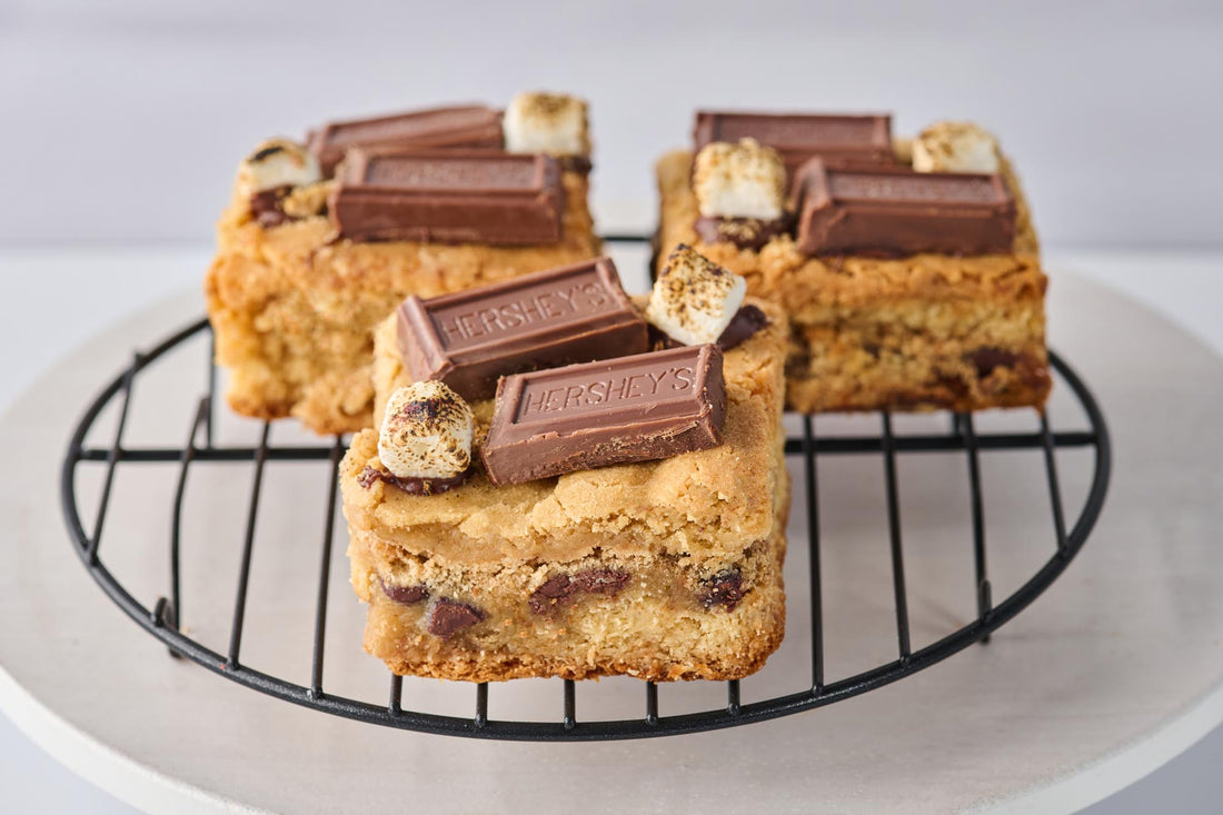 S'mores Blondie with rich buttery base, pockets of melted chocolate and gooey marshmallow, topped with graham cracker crumbs, toasted marshmallows, and Hershey's chocolate for a decadent s'mores treat.
